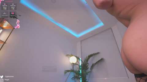 Media: Video of a topless woman with large breasts, standing in a modern, dimly lit room with white walls and blue LED lights. A fern plant and a lamp are in the background.