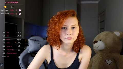 Media: Video of a young woman with curly red hair, fair skin, wearing a black tank top, seated in a dimly lit room with a grey chair and teddy bear.