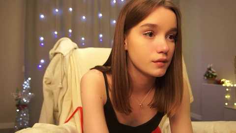 Media: Video of a young Caucasian girl with straight, shoulder-length brown hair, wearing a black tank top and red cardigan, sitting on a beige sofa, looking thoughtful. Background features festive string lights and a Christmas tree.