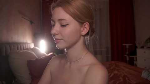 Media: Video of a young Caucasian woman with fair skin and light brown hair, wearing minimal makeup, sitting topless in a dimly lit bedroom, with red curtains, a white dresser, and a bed with brown bedding in the background.