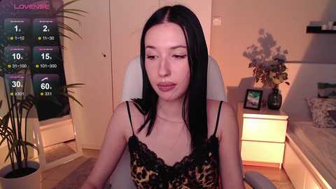 Media: A video of a fair-skinned woman with straight black hair, wearing a black lace bra, seated in a modern, dimly-lit room with a digital weather overlay.