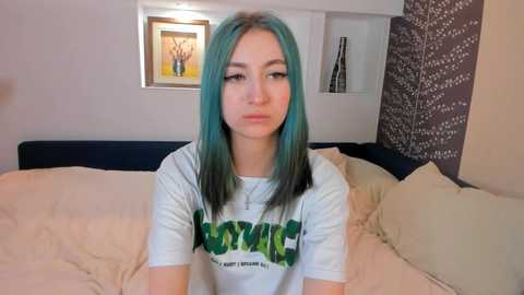 Media: Video of a young woman with teal hair, light skin, and a neutral expression, wearing a white t-shirt with green text, sitting on a bed with beige sheets and pillows, in a cozy, modern bedroom with wall art and patterned wallpaper.