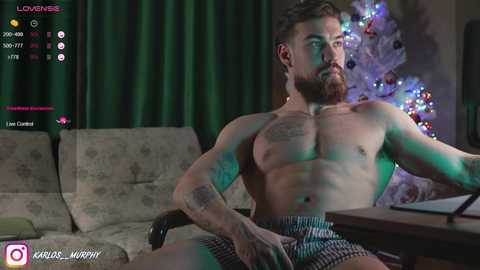 Media: Video of a muscular, shirtless man with tattoos, sitting in a living room with a Christmas tree. He wears checkered underwear, with green and purple lighting.