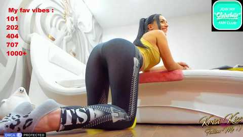 Media: Video of a woman with long black hair, wearing a yellow sports bra and black leggings, bending over a white bed, smiling. Background features a white room with a hanging plant.