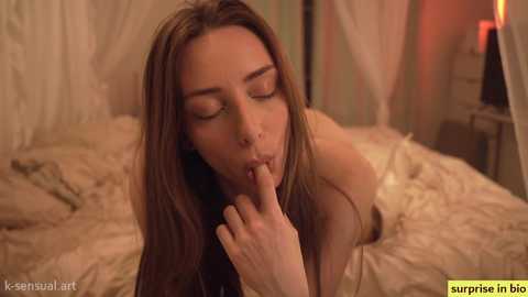 Media: A video of a topless young woman with long brown hair, performing a sensual act on a bed, in a dimly-lit, cozy bedroom.