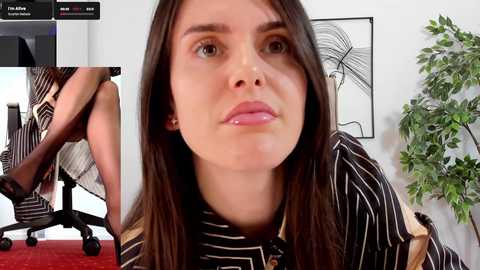 Media: Video of a young woman with straight brown hair, wearing a striped dress, sitting on a chair in a modern office with a green plant and abstract art on the wall.