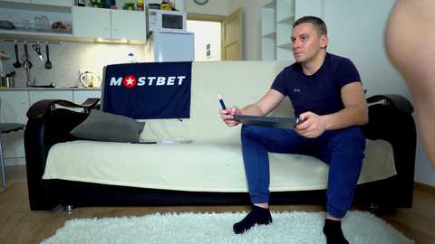 Media: A video of a muscular, shirtless man in blue pants and socks, holding a clipboard and a pen, sitting on a beige couch in a modern, minimalist living room.