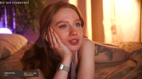 Media: Video of a fair-skinned woman with long red hair, wearing a gray tank top, lying on a bed, looking thoughtful, with a tattoo on her arm, in a warmly lit room.