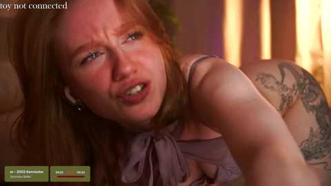 Media: A video of a red-haired, fair-skinned woman with a distressed expression, wearing a purple top and visible tattoos. Background features warm, dim lighting and a blurred, indistinct room.