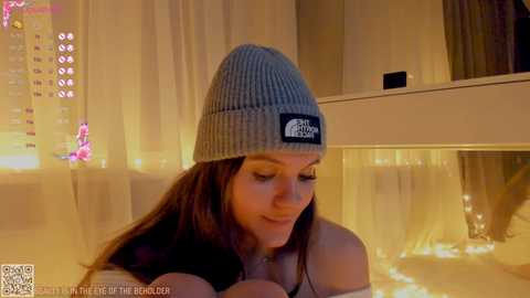 Media: Video of a smiling woman with long brown hair, wearing a grey beanie and off-shoulder top, standing in a warmly lit room with string lights.