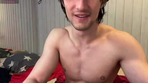 Media: Video of a young, shirtless white male with short, dark hair, smiling, in a casual, dimly lit room with wooden paneling and a bed with red and gray bedding.