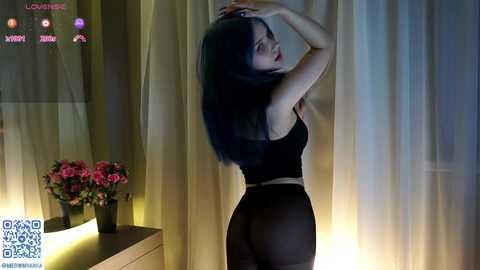 Video of a slender Asian woman with long black hair, wearing a black crop top and sheer black pants, posing seductively against a backdrop of sheer white curtains.