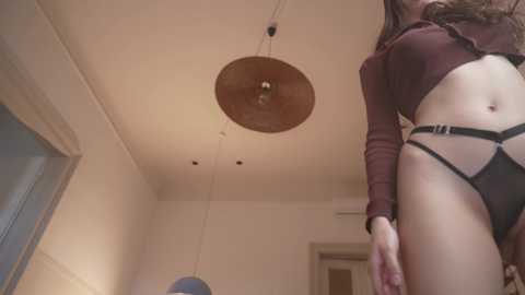 Media: Video of a slender woman with long brown hair, wearing a maroon crop top and black strappy lingerie, standing in a modern, dimly lit room with beige walls and a circular wooden chandelier.