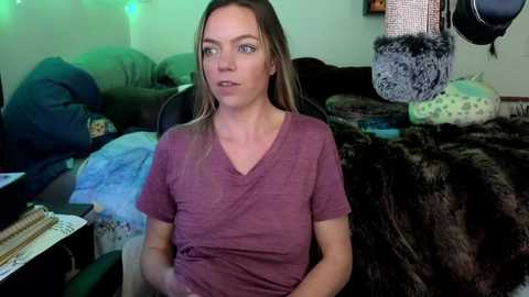 Media: Video of a Caucasian woman with light skin, long blonde hair, and blue eyes, wearing a purple V-neck t-shirt, sitting on a black chair in a cluttered living room with greenish lights, fur blankets, and pillows.