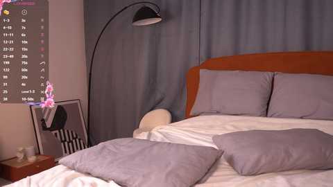 Media: Video of a minimalist bedroom with a modern design: a queen-sized bed with white linens and gray pillows, a large black lamp, and a gray curtain backdrop.