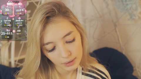 Media: Video of a young blonde woman with closed eyes, soft skin, and a relaxed expression, wearing a striped shirt. Background features a cozy, blurred room with a bed and a clock displaying 25:11.