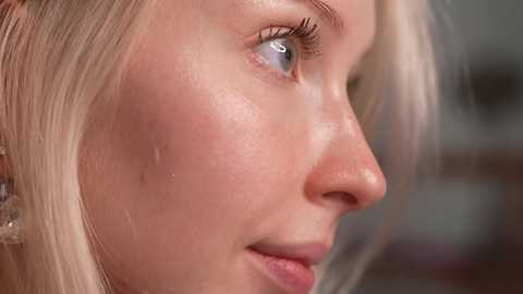 Close-up video of a fair-skinned, blonde woman with a shiny forehead, possibly from sweat or oil, wearing minimal makeup, with blue eyes and a soft smile.