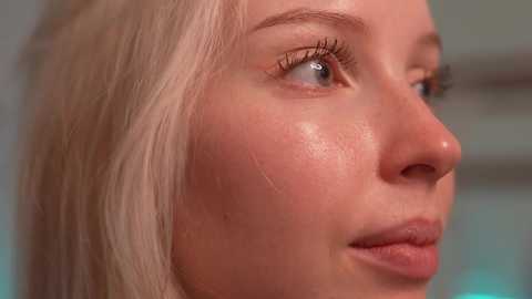 Media: Close-up video of a fair-skinned woman with blonde hair, showing her profile. Her skin has a shiny, smooth texture, with visible pores and a slight blush on her cheek. The background is blurred, highlighting her features.