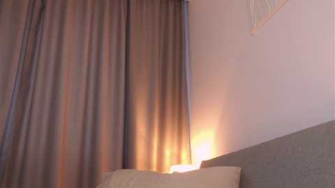 Media: Video of a cozy bedroom corner with a grey headboard, beige pillows, and a soft, warm light glowing from behind a sheer curtain, casting a gentle, golden hue on the walls.