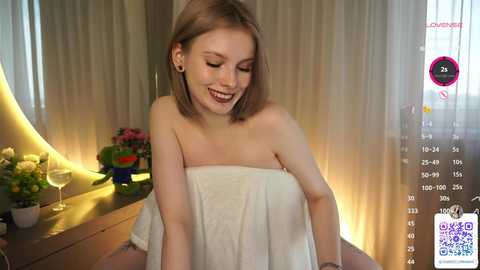 Media: Video of a young Caucasian woman with light skin and straight, shoulder-length brown hair, smiling while wrapped in a white towel, sitting on a wooden table. The background features a floral arrangement and sheer curtains.