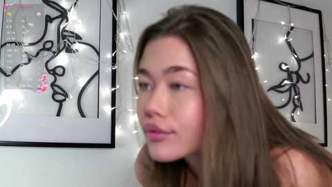 Media: A video of a young woman with fair skin and long brown hair, looking contemplative. She is indoors with modern, minimalist art and string lights in the background.