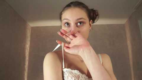 Media: Video of a young woman with fair skin and dark hair in a bun, wearing a white lace bra, touching her nose with her fingers. She stands in a beige-tiled bathroom with a mirror and soft lighting.