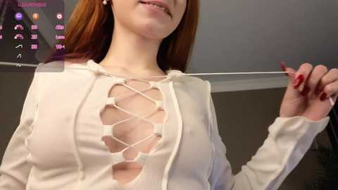 Video of a fair-skinned woman with red hair, wearing a revealing white top with cutouts, holding a lanyard. Background shows a digital interface with social media icons.