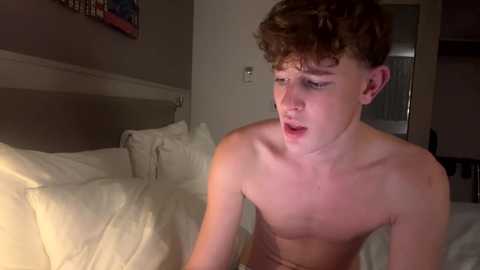 Media: Video of a young, fair-skinned, curly-haired, shirtless teenage boy sitting on a bed with white sheets, looking surprised, in a dimly lit room.