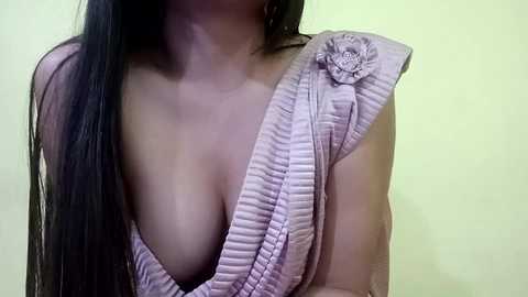 Media: Video of a woman with long, straight black hair, wearing a pink, ruffled off-shoulder top with a floral embellishment. The background is a plain, light green wall. Her breasts are partially visible under the low-cut neckline.