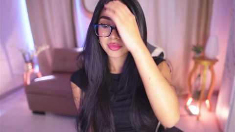 Media: Video of a woman with long black hair, glasses, and red lipstick, covering her face with her hand, sitting in a modern living room with beige furniture and soft lighting.