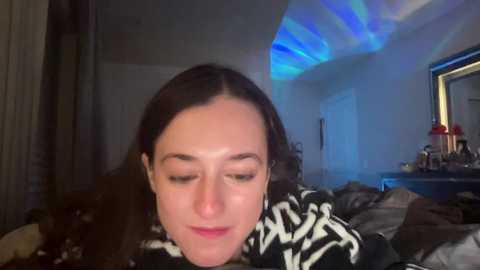 Media: Video of a young woman with long brown hair, wearing a black and white sweater, lying on a messy bed, under blue LED lights in a dimly lit bedroom.