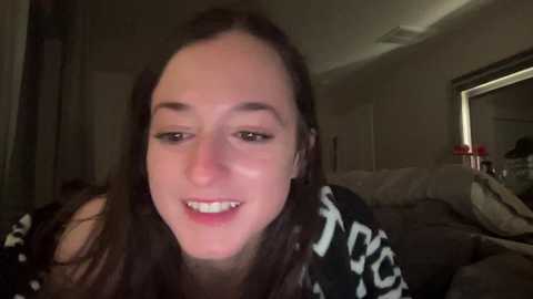 Media: A video of a smiling, young woman with fair skin and long brown hair, wearing a black and white patterned top. She is indoors, in a dimly lit room with a bed and a mirror visible in the background.