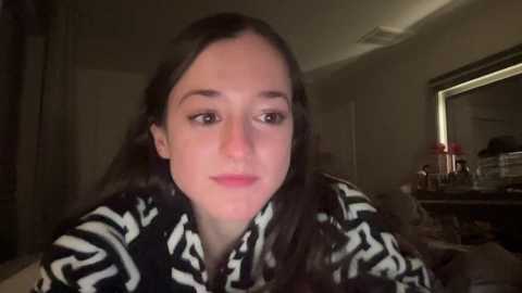 Media: A video of a young woman with fair skin, long dark hair, and brown eyes, wearing a black and white patterned shirt, in a dimly lit room with a mirror and cluttered background.