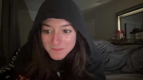 Media: Video of a young woman with long, dark hair and fair skin, wearing a black hooded sweatshirt, looking into the camera. The background shows a dimly lit bedroom with messy bedding and a red gift box on a nightstand.