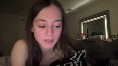 Media: Video of a young woman with long dark hair, fair skin, and closed eyes, wearing a black and white patterned top, sitting in a dimly lit bedroom with a mirror and cluttered nightstand.