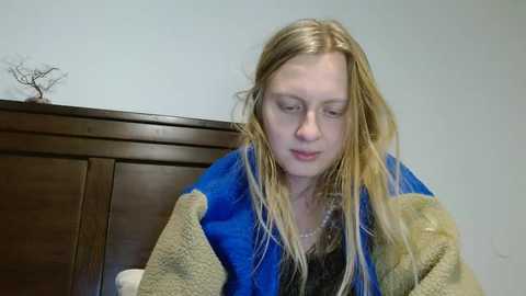 Media: A video of a Caucasian woman with long blonde hair, wearing a blue and beige blanket, sitting on a wooden headboard against a plain light blue wall.