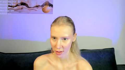 Media: Video of a fair-skinned woman with blonde hair, wearing nothing, sitting on a black couch against a purple wall. A poster with a beach scene is visible in the background.