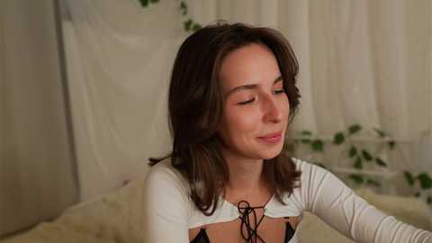 Media: Video of a smiling Caucasian woman with medium-length brown hair, wearing a white, long-sleeved top with a black lace-up front, in a softly lit room with white curtains and potted plants.