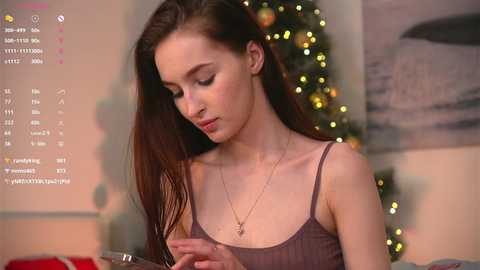 A video of a young woman with long brown hair, wearing a gray tank top, standing in front of a Christmas tree with glowing lights.