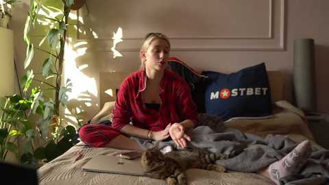 Media: Video of a blonde woman in a red robe sitting cross-legged on a bed with a cat. The room is bright, with sunlight streaming in, and has a cozy, relaxed atmosphere.