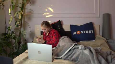 Media: Video of a woman with blonde hair in a red plaid shirt, reading a book on a laptop, wrapped in a gray blanket on a bed with a \"MOSTBET\" pillow, surrounded by plants.