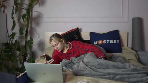 Media: Video of a blonde woman in red plaid pajamas, reading on a bed with a laptop and gray blanket. Background includes a plant and a \"MOSTBET\" pillow.