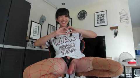 Media: Video of a smiling, slender Asian woman with black hair, wearing a \"I AM BANG YANKY\" t-shirt, fishnet stockings, and a visible penis. She sits on a black chair in a modern living room with minimalistic decor.