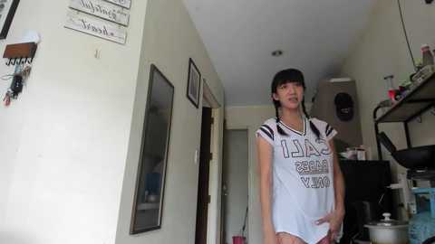 Media: Video of a young Asian woman with long black hair, wearing a white soccer jersey with black text, standing in a cluttered, dimly lit room with a doorway and shelves visible.