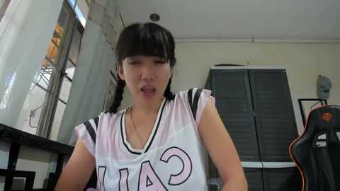 Media: Video of an East Asian woman with long black hair in braids, wearing a white and black \"CAPS\" jersey, sitting at a desk in a cluttered room with a window and a gaming chair.