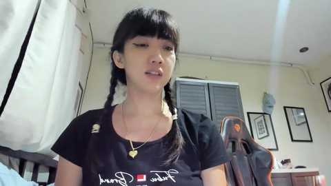 Media: Video of a young Asian woman with straight black hair in braids, wearing a black t-shirt with a Japanese script and gold heart pendant. She's in a simple bedroom with white walls, a window, and a chair.