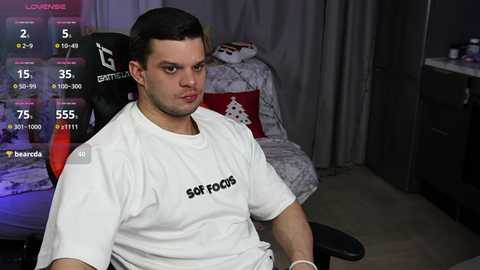 Video of a muscular, light-skinned man with short dark hair, wearing a white \"So Fatt\" T-shirt, seated in a gaming chair in a dimly lit room with gaming setup.