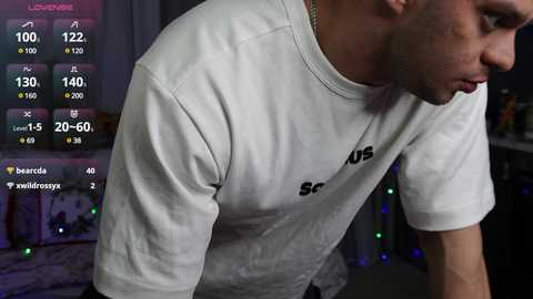 Video of a young man in a white T-shirt, bending over, taken indoors. Background shows a colorful, dimly-lit room with gaming equipment and a TV displaying stats and graphics.