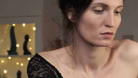 Media: Video of a woman with fair skin and dark hair in a messy bun, wearing a black lace dress, looking contemplative. Background features a blurred shelf with yellow lights and indistinct objects.