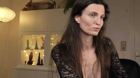 Media: Video of a woman with long brown hair, wearing a sheer black lace top, standing in a cozy, warmly lit room with a bookshelf displaying sex toys.